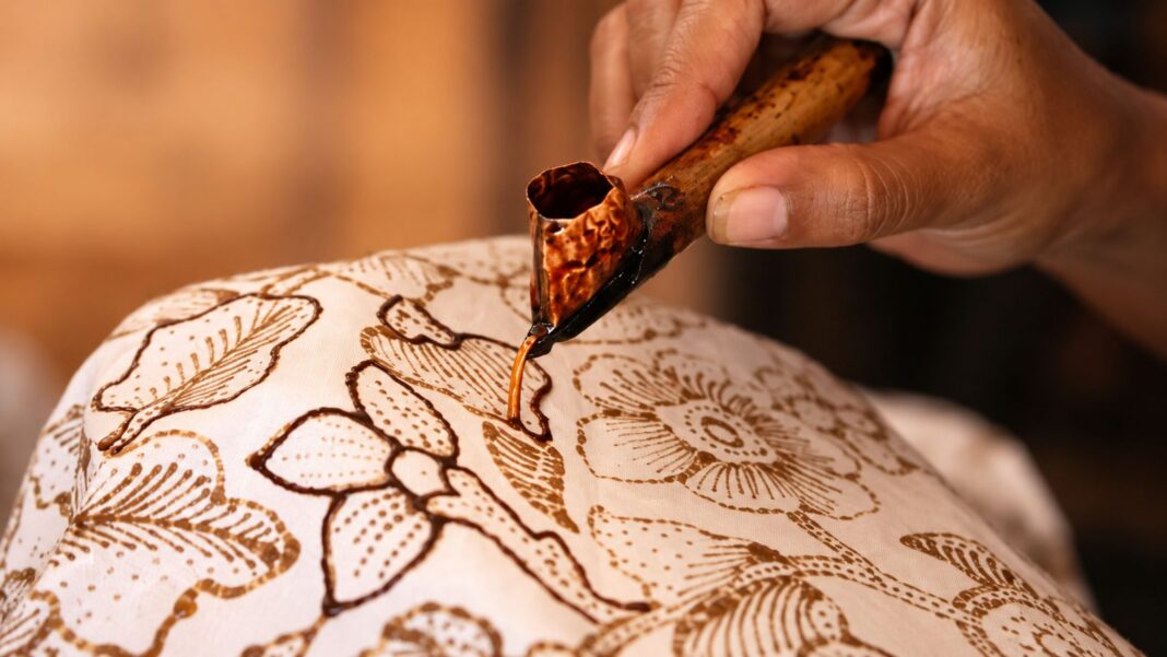 Proses batik tulis: tangan perajin menggoreskan canting berisi malam panas di atas kain bermotif bunga